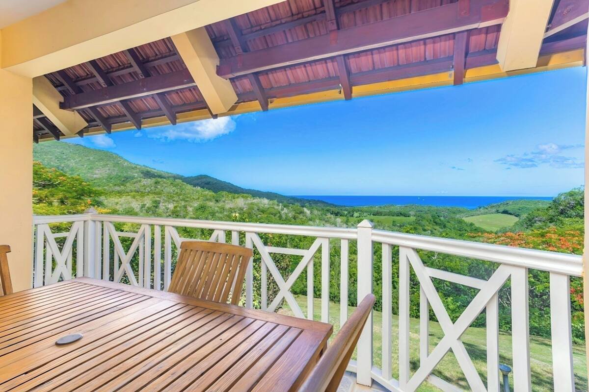 Ein Holztisch und -stühle auf einem Balkon mit weißer Brüstung, mit Blick auf eine üppige grüne Landschaft und das Meer.