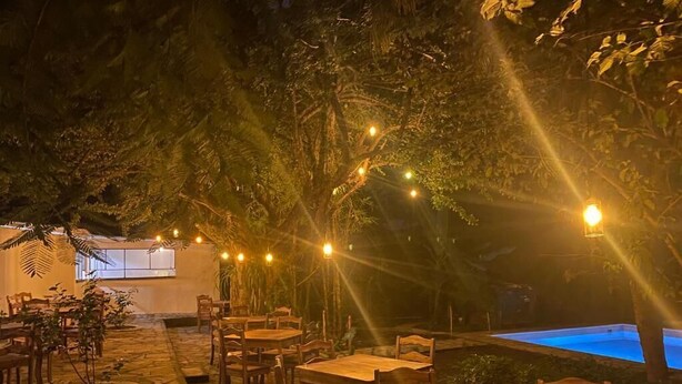 Un espace extérieur aménagé pour les repas, avec un sol en pierre, des tables et des chaises en bois, des guirlandes lumineuses et une piscine en arrière-plan.