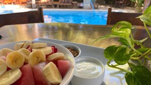 Un saladier rempli de salade de fruits contenant des bananes et de la pastèque, un petit bol de yaourt et une plante sur une table près d’une piscine.