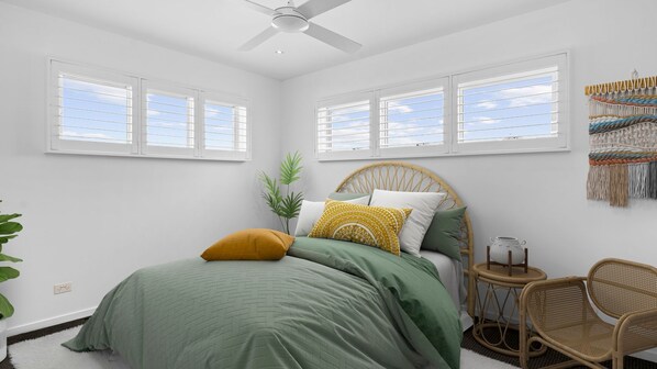 A bedroom with a bed, two windows, a ceiling fan, a rattan chair, and a wall hanging.