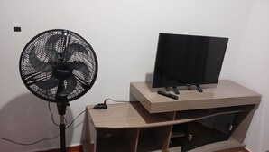 A room with a wooden TV stand holding a flat-screen TV and a remote control. A black standing fan is positioned to the left of the TV stand. - Beautiful and cozy house, spacious and close to the center (Boa Esperança Toledo)
