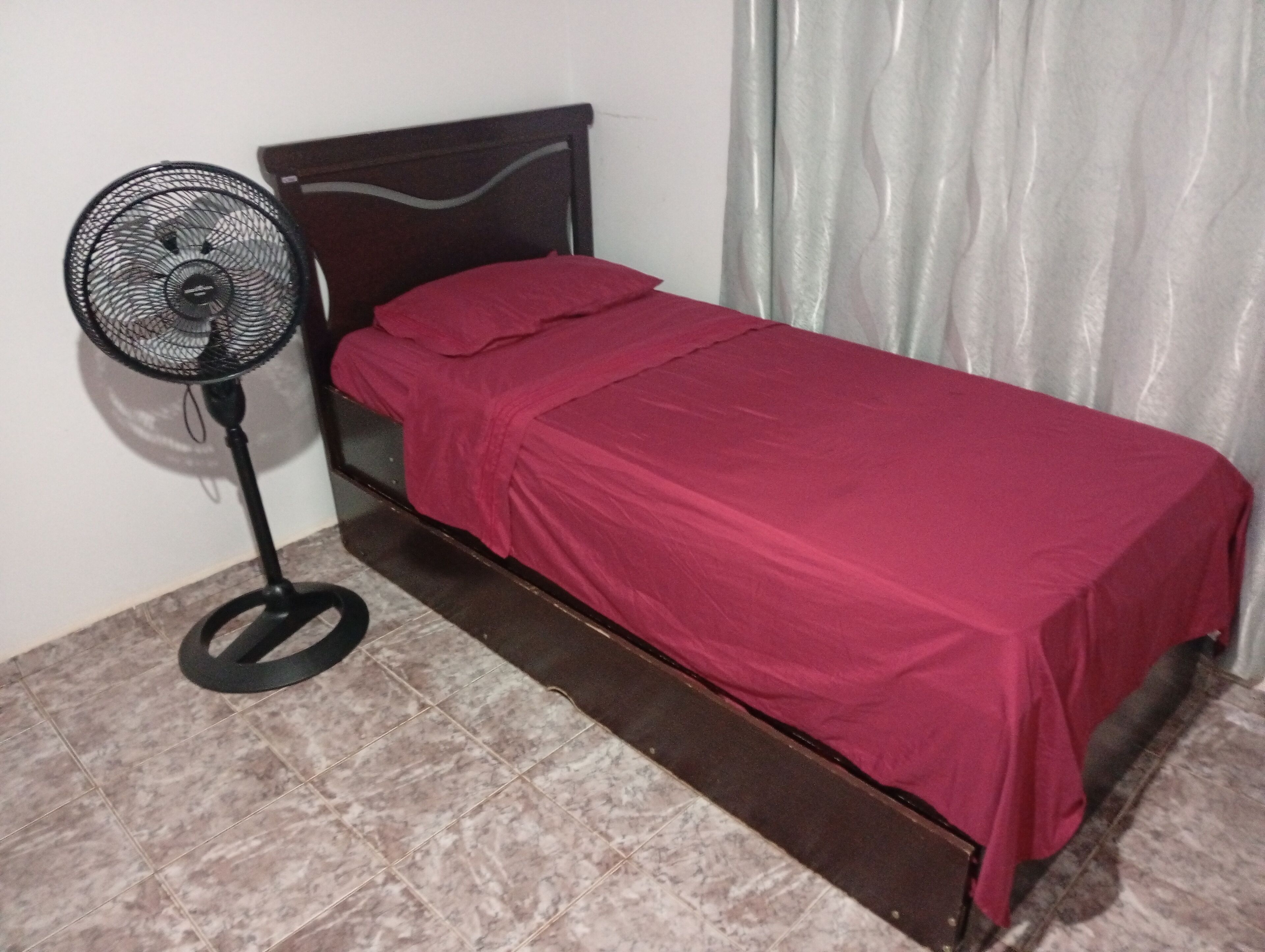 A single bed with a red bedspread, a standing fan, and a curtain.