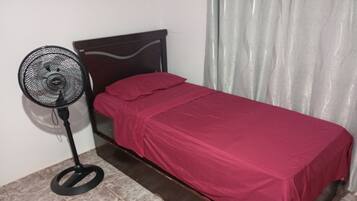 A single bed with a red bedspread, a standing fan, and a curtain.