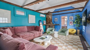 Un soggiorno con un divano angolare rosa, un tavolino da caffè bianco e un tappeto a scacchi. C'è un caminetto in mattoni, una poltrona verde e un pouf giallo. Le pareti sono dipinte di blu e ci sono quadri appesi.