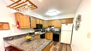 A kitchen with wooden cabinets, granite countertops, a central island with stools, a microwave, a refrigerator, and a hanging light fixture.