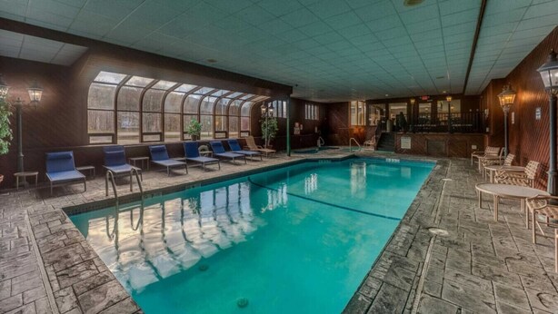 An indoor swimming pool with lounge chairs, a bar area, and a view through a glass enclosure.