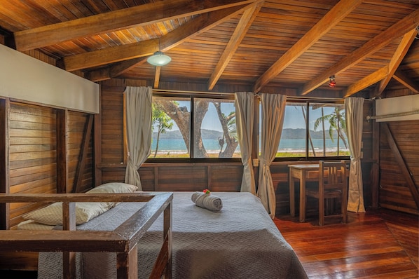 A wooden room with a bed, a table, and a view of the beach.