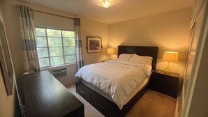 Une chambre à coucher avec un lit, deux tables de chevet avec des lampes, une fenêtre avec des rideaux, un radiateur et un tableau au mur.