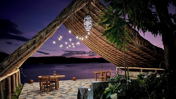 An outdoor dining area with a thatched roof, string lights, and a view of the water at dusk.
