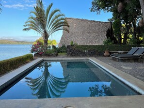 Ein Pool mit klarem Wasser, das Palmen und eine Steinmauer spiegelt, umgeben von Liegestühlen und Blick auf das Meer.