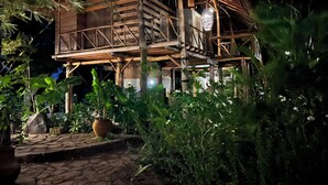 A wooden house with a thatched roof, surrounded by lush greenery.