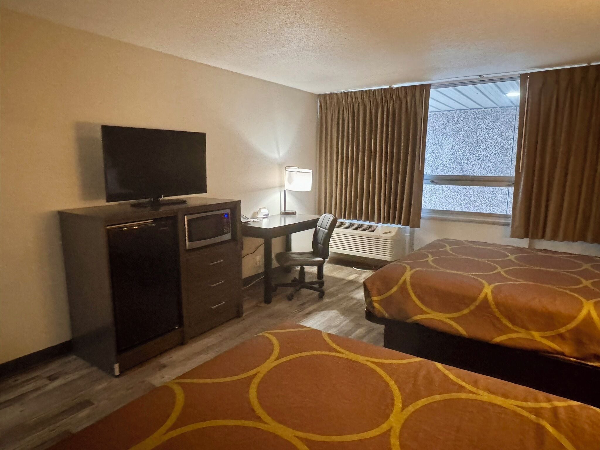 A hotel room with a desk, a television, a chair, two beds, a heater, and a window with curtains.