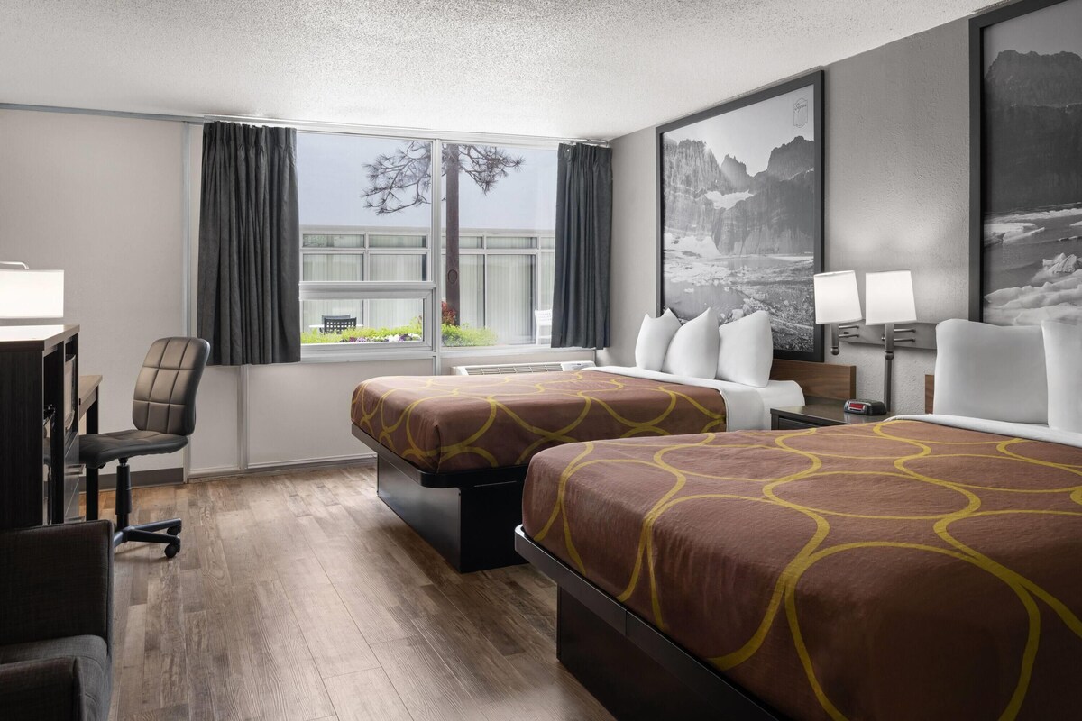 A hotel room with two beds, a desk, a chair, a window with curtains, and a view of greenery.