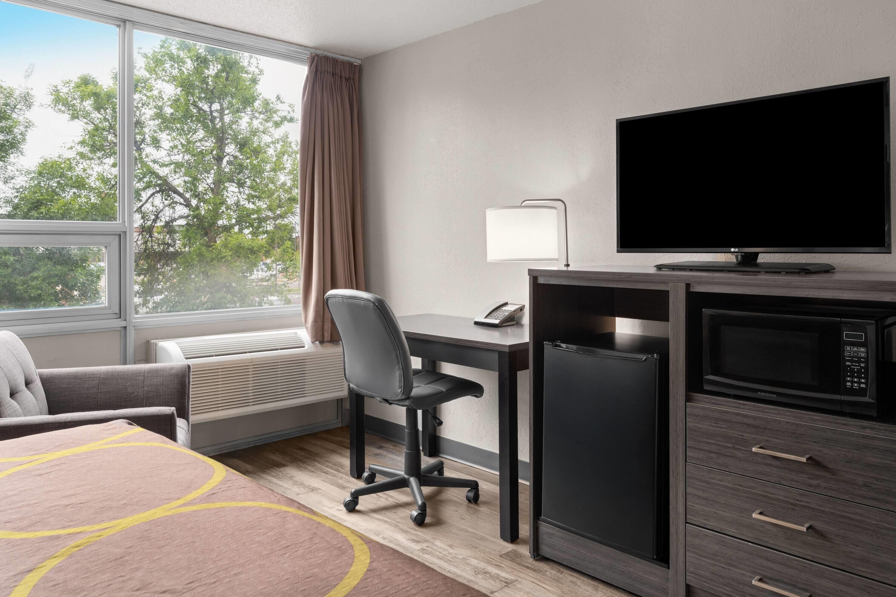 Quarto de hotel com escrivaninha, cadeira, televisão, micro-ondas, frigorífico e uma janela grande com cortinas.