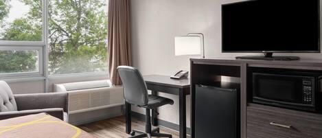 A hotel room with a desk, chair, television, microwave, refrigerator, and a large window with curtains.