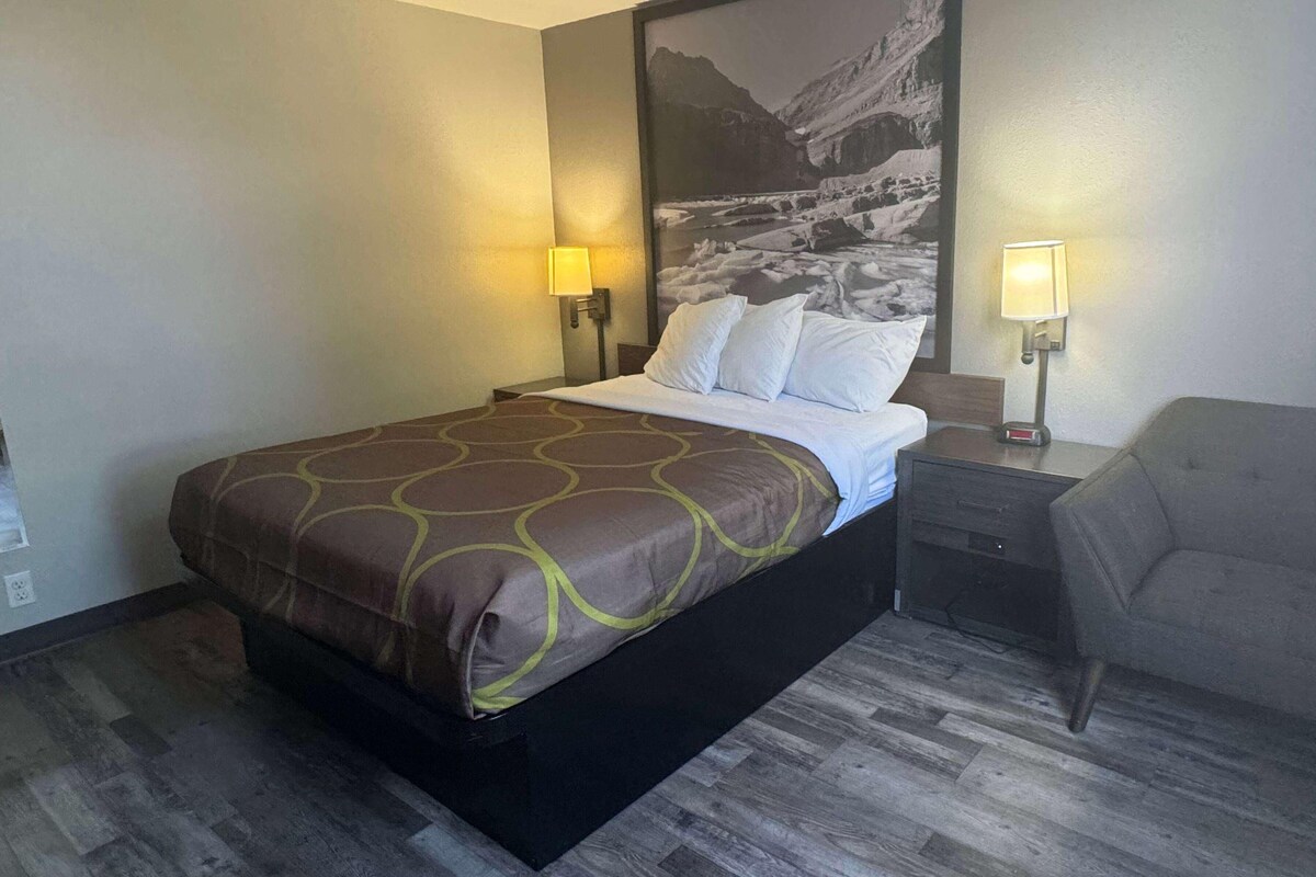 Une chambre d’hôtel avec un lit, deux lampes de chevet, une table de chevet, une chaise et un grand tableau mural.