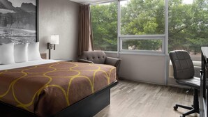 A hotel room with a bed, a desk, a chair, a window with a view of trees, and a wall-mounted lamp. - Super 8 By Wyndham Great Falls City Center (Great Falls)