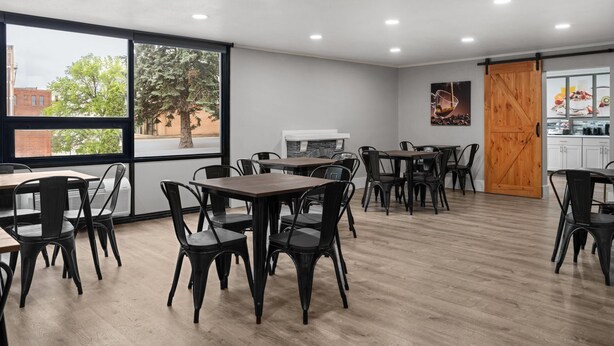 A modern dining area with tables and chairs, large windows with a view of trees and buildings, and a kitchen area visible through a sliding door. - Super 8 By Wyndham Great Falls City Center (Great Falls)
