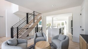Una sala de estar moderna con una escalera, sillones grises, una mesa de centro redonda de madera y una cocina al fondo.