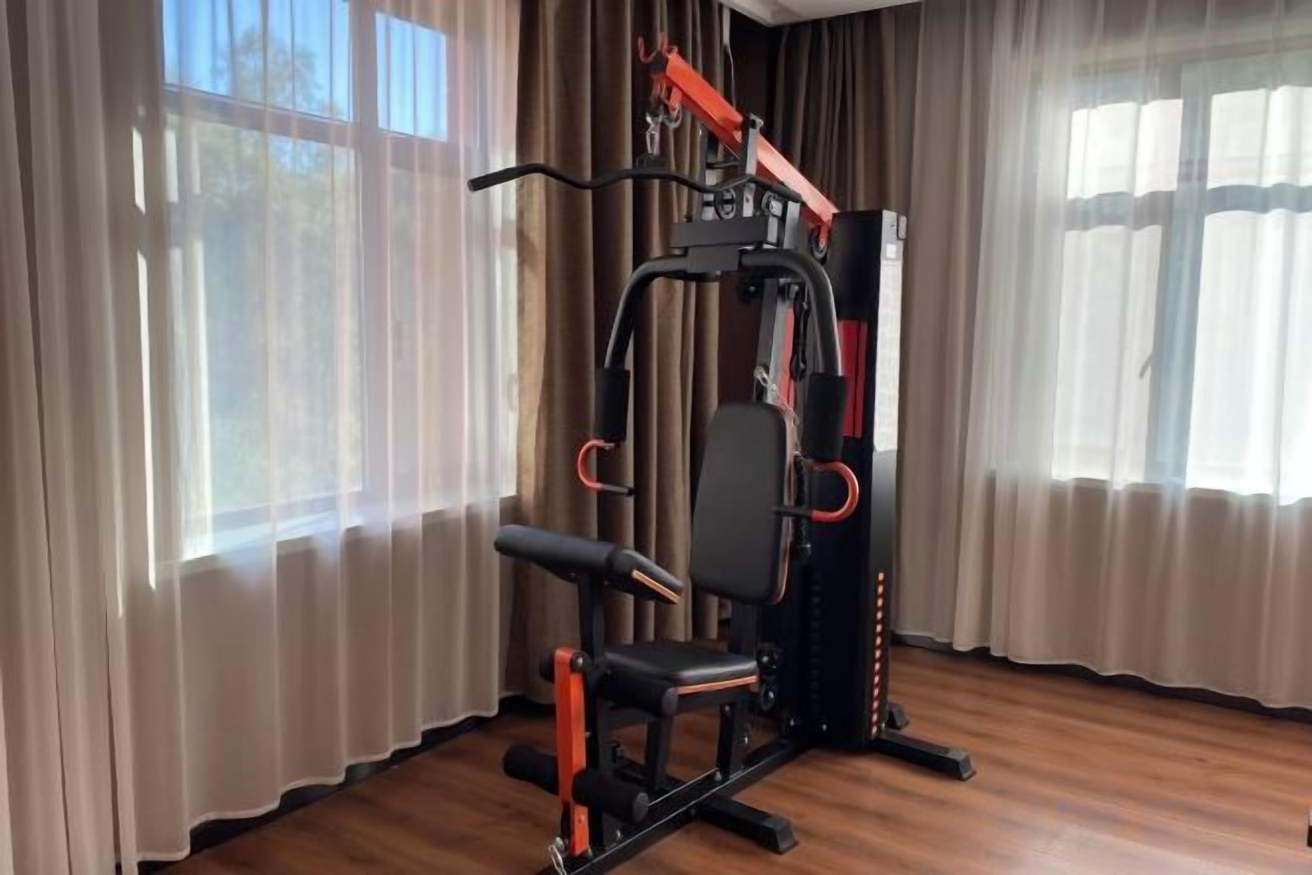 Een fitnessruimte met een krachttrainingapparaat en een bank, gelegen in een ruimte met grote ramen en gordijnen.