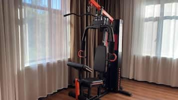 Een fitnessruimte met een krachttrainingapparaat en een bank, gelegen in een ruimte met grote ramen en gordijnen.