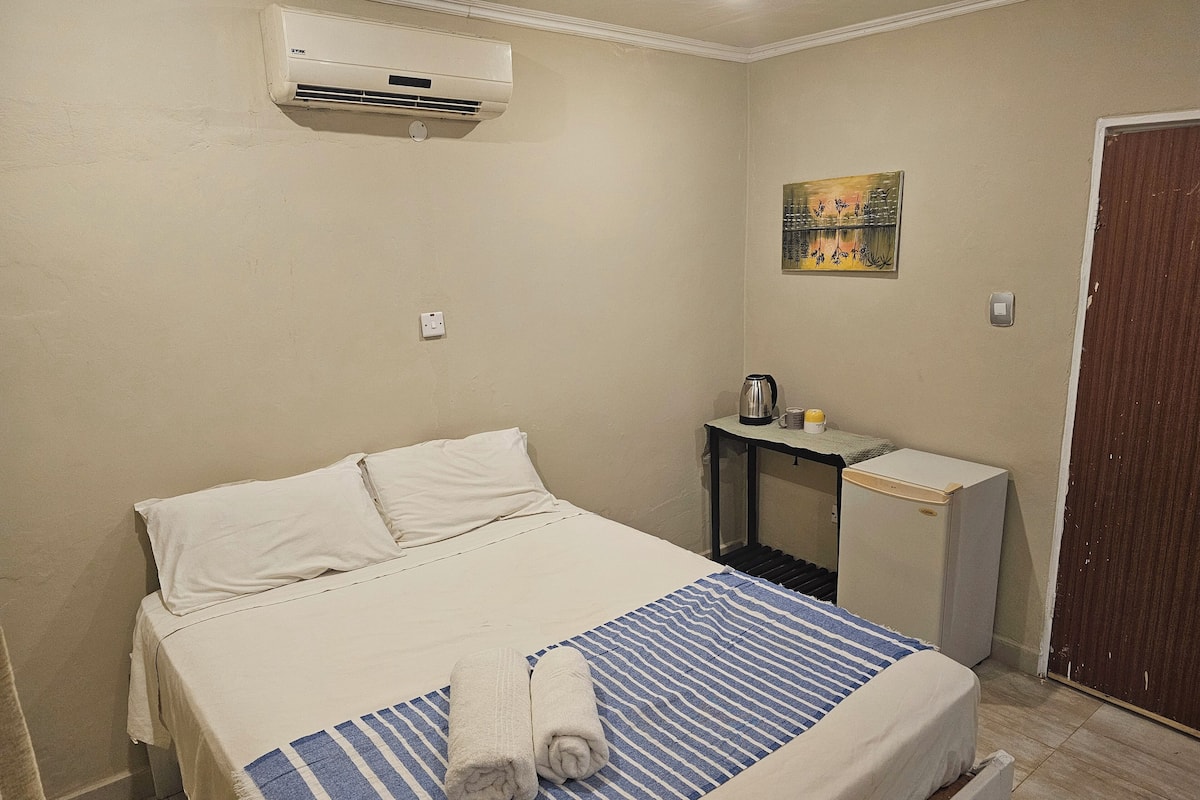 Un dormitorio con cama, mesita de noche con tetera y taza, aire acondicionado y un cuadro en la pared.