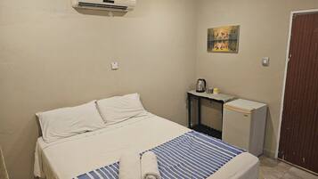 A bedroom with a bed, a bedside table with a kettle and mug, an air conditioner, and a painting on the wall.