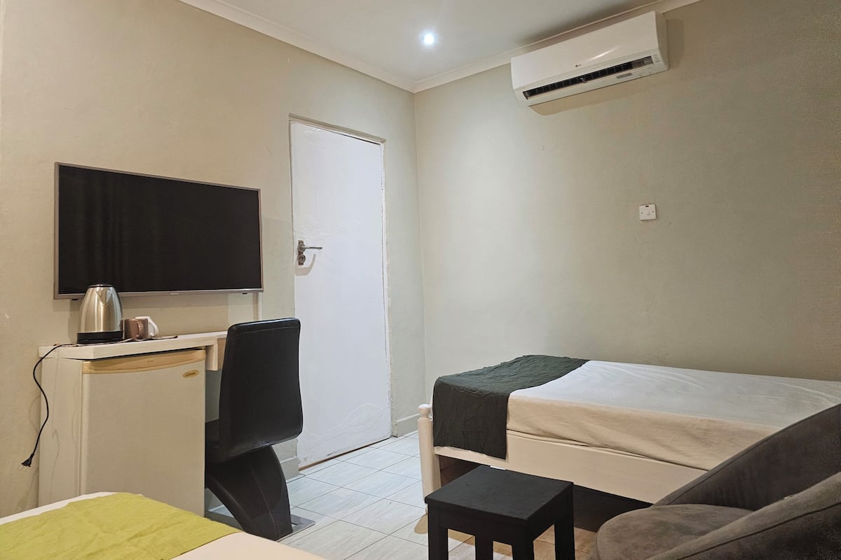 Una habitación pequeña y limpia con una cama, un escritorio con silla, televisión y aire acondicionado.