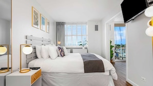 Un dormitorio moderno con cama, mesitas de noche, televisor y ventana con vista a barcos.