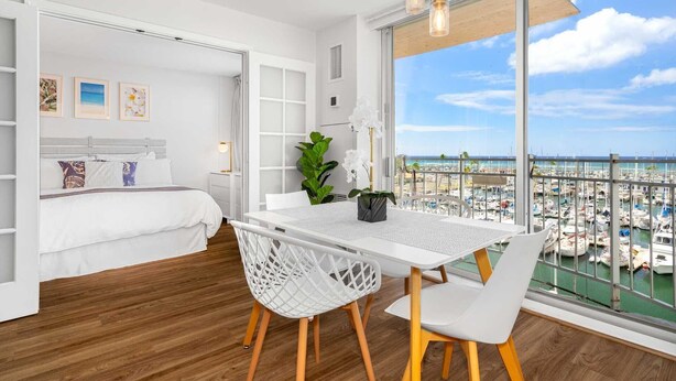 Un dormitorio moderno con un ventanal que da a un puerto deportivo, un área de comedor blanca con una mesa y sillas, y una cama con ropa de cama blanca.