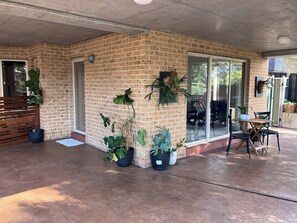 Ein überdachter Patio-Bereich mit Backsteinmauern, Topfpflanzen, einem Tisch mit Stühlen und einer Glasschiebetür, die zu einem Außenbereich führt.