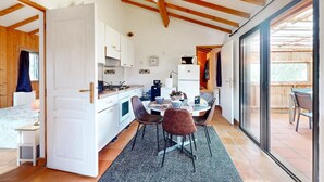 Cocina compacta con gabinetes blancos, estufa y refrigerador. Hay una zona de comedor con mesa y sillas, y un gran ventanal corredera que da a un patio exterior.