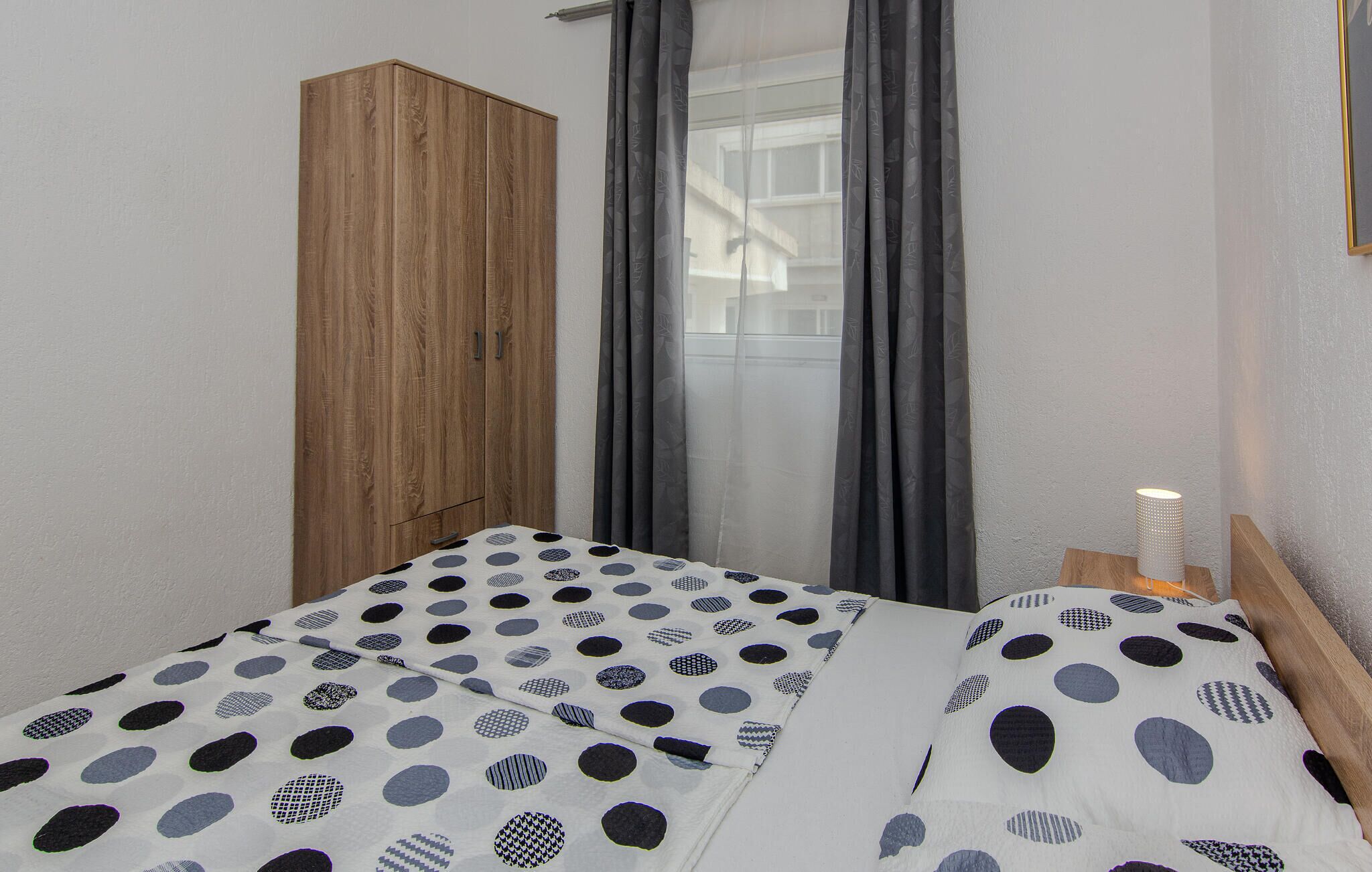 Une chambre à coucher avec un lit recouvert d’une courtepointe à motifs à pois et géométriques, une armoire en bois et une fenêtre ornée de rideaux gris.