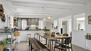 En rummelig spiseplads med et langt træbord, sorte stole og en bænk. Et køkken er synligt i baggrunden med moderne hårde hvidevarer og en hængende lampe.