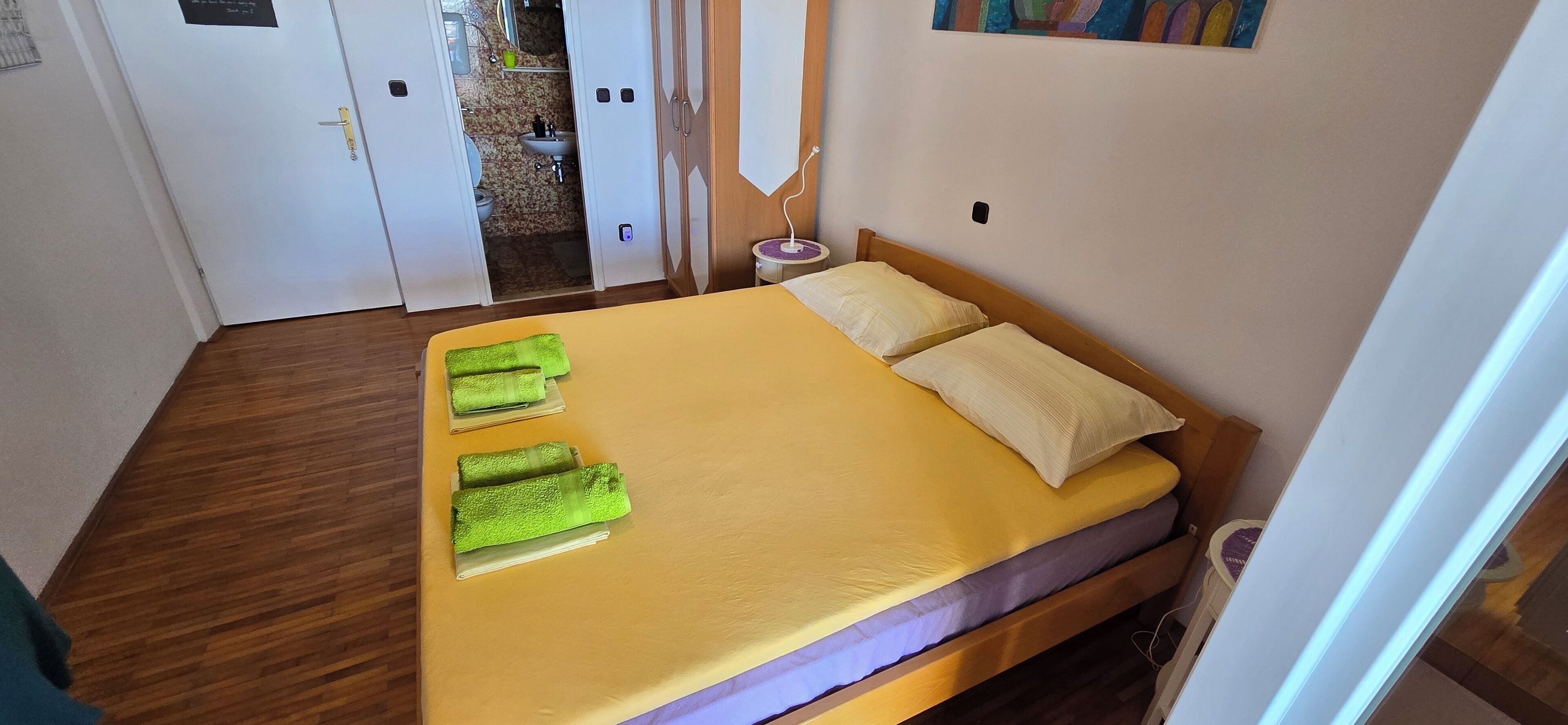 A small, neatly made bed with a yellow mattress and two pillows, a wooden floor, and a door leading to a bathroom.