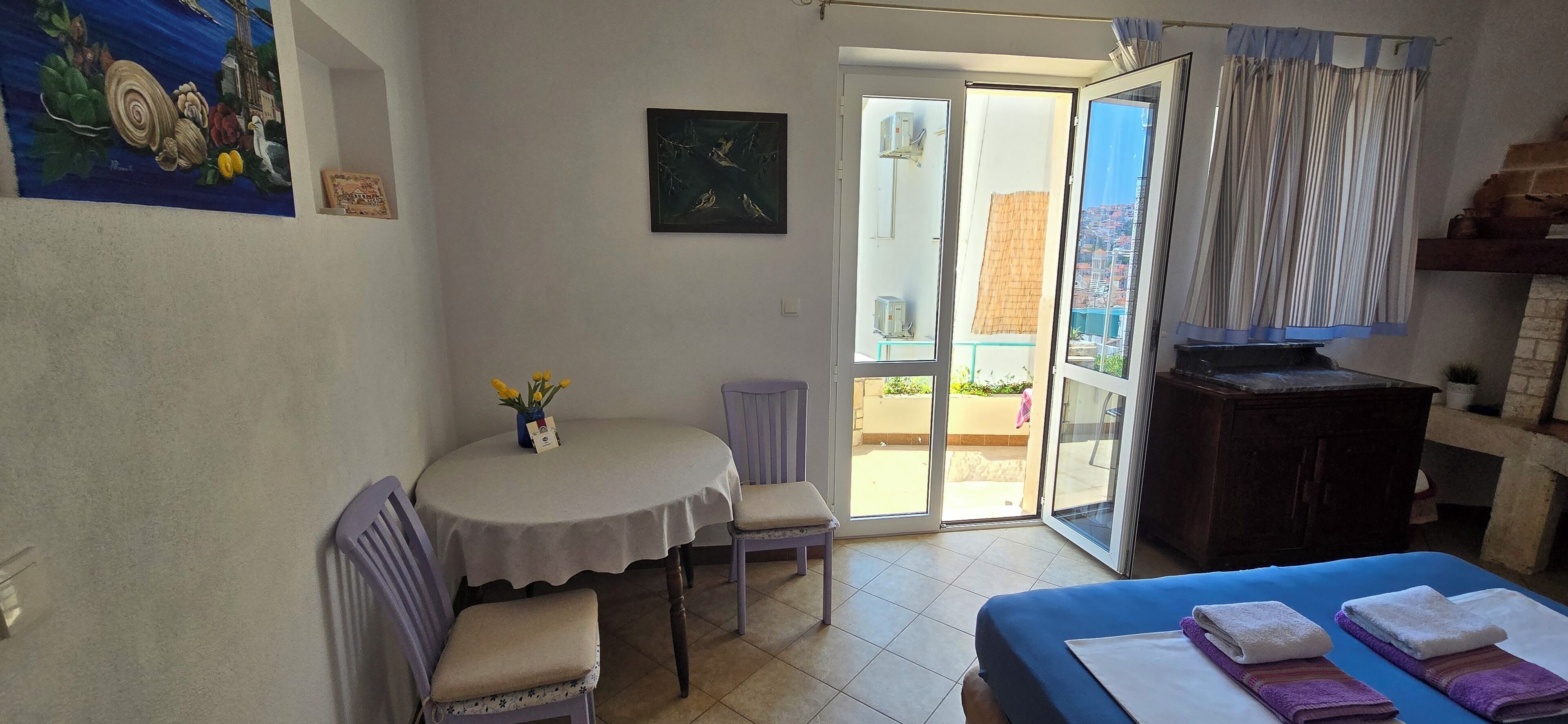 A room with a bed, a table with chairs, a painting on the wall, and a view through the open door.