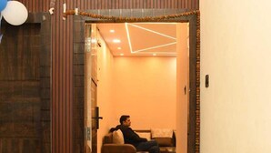 A hotel entrance with a wooden door, a person sitting on a couch inside, and a geometric light fixture on the ceiling. - Abhivadya Gange  (Varanasi)