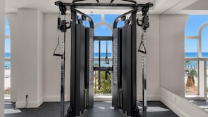 Un gimnasio con barra de dominadas y máquinas de pesas, amplias ventanas y vista a la playa.