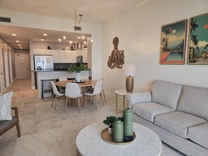 Uma sala de estar moderna com um sofá cinza, uma área de refeições com uma mesa redonda e cadeiras, uma cozinha ao fundo e obras de arte na parede.