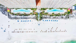 Vista aérea de un resort con dos piscinas, una zona de playa con tumbonas y un paseo cubierto.