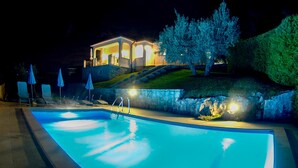 A nighttime swimming pool area with lounge chairs and umbrellas, a house with illuminated windows, and a landscaped hill with trees. - Gorgeous home in Vizinada (Vizinada)