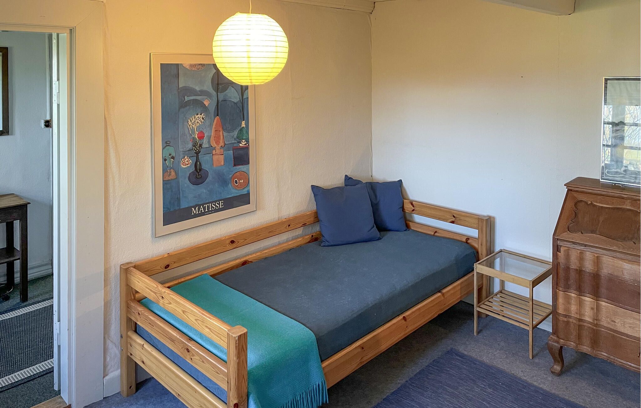 Ein kleiner, gemütlicher Raum mit einem hölzernen Bett, einem blauen Kissen, einem gerahmten Kunstwerk an der Wand und einer Holzkommode.