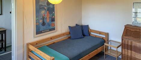 Ein kleiner, gemütlicher Raum mit einem hölzernen Bett, einem blauen Kissen, einem gerahmten Kunstwerk an der Wand und einer Holzkommode.