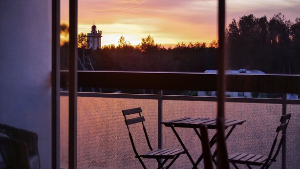 Un balcón con dos sillas de madera y una mesa, con vistas a una masa de agua y un edificio distante al atardecer.