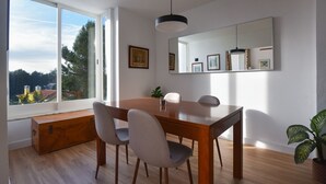 Un comedor con una mesa de madera, cuatro sillas, un banco y un amplio ventanal con vistas a árboles y edificios.