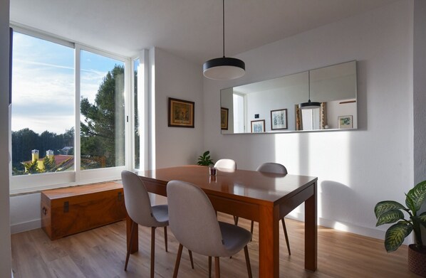 A dining area with a wooden table, four chairs, a bench, and a large window offering a view of trees and buildings.