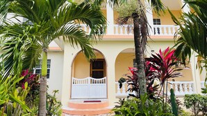 Un edificio a due piani con un balcone bianco, un ingresso piastrellato e rigogliosa vegetazione tropicale.