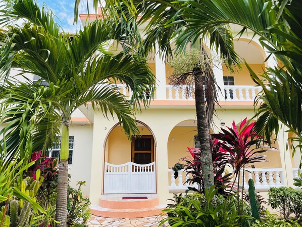 A two-story building with a white balcony, a tiled entrance, and lush tropical plants.