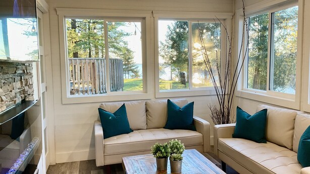 Ein gemütliches Wohnzimmer mit einem beigen Sofa, blaustichigen Kissen und einem Holztisch mit Pflanzen. Große Fenster bieten einen Ausblick ins Freie.
