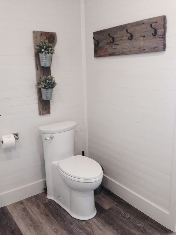 A small, white bathroom with a toilet, a wooden wall-mounted shelf with potted plants, and a rustic wall decoration. - Riviera - A gathering spot (Lansdowne)